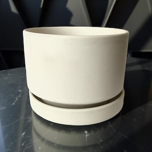 May include: A cream-colored ceramic planter with a matching saucer. The cylindrical pot has a smooth, matte finish and sits on a round base. The planter is designed for indoor or outdoor use.