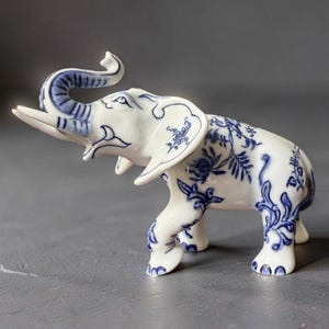 Op de afbeelding: Een wit keramieken olifantenbeeldje met gedetailleerde blauwe bloemen- en rankpatronen over het hele lichaam. De olifant is afgebeeld met de slurf omhoog en de slagtanden naar voren gericht, met een gladde en glanzende afwerking.