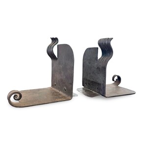 May include: A pair of handcrafted, dark metal bookends. Each bookend features a flat base with a curled end and a vertical support with a decorative, wavy design. The metal has a rustic, aged appearance.