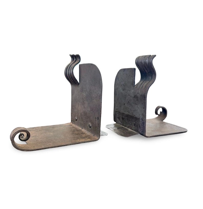 May include: A pair of handcrafted, dark metal bookends. Each bookend features a flat base with a curled end and a vertical support with a decorative, wavy design. The metal has a rustic, aged appearance.