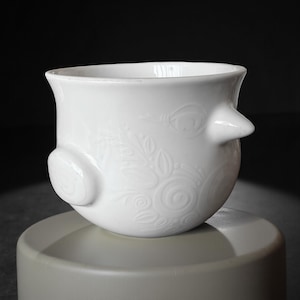 Op de afbeelding: Een witte keramische kom met een vogelachtig ontwerp. De kom heeft een afgeronde body met een uitlopende rand en twee uitstekende elementen die lijken op een snavel en een vleugel. Er zijn ingewikkelde bloempatronen in reliëf op het oppervlak.