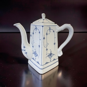 Op de afbeelding: Witte porseleinen koffiepot met een vierkant lichaam en deksel, versierd met blauwe bloemen- en wijnstokpatronen. Het heeft een gebogen tuit en een handvat. Het ontwerp bevat een blauwe rand. Een klassiek stuk serviesgoed.