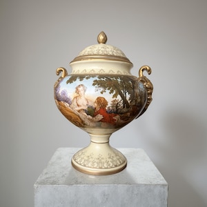 Op de afbeelding: Crèmekleurige keramische urn met deksel, versierd met een geschilderde pastorale scène. Gouden accenten sieren de handvatten, rand en knop. De urn heeft een voetstuk. De scène toont figuren in een landschap.