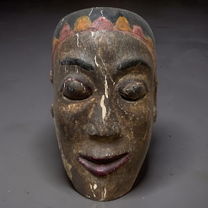 May include: A hand-carved wooden mask featuring a dark brown weathered finish, prominent almond-shaped eyes, and a defined nose. The forehead is adorned with a scalloped pattern in yellow and red, while the lips are painted a deep reddish-purple.