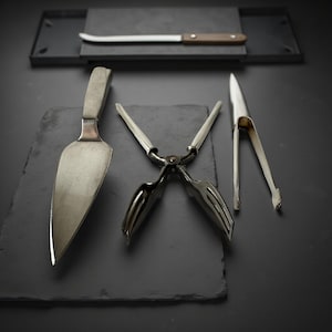 May include: A collection of stainless steel serving utensils, including a cake server, tongs, and a knife with a wooden handle. The items are arranged on a dark gray surface.