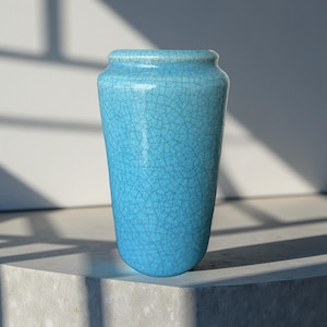 Könnte beinhalten: Eine hohe, türkisfarbene Keramikvase mit Craquelé-Glasur. Die Vase hat einen abgerundeten Boden und einen leicht ausgestellten Rand. Sie steht auf einer Betonoberfläche, mit Schatten von einem Fenster im Hintergrund.