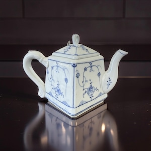Könnte beinhalten: Eine weiße Porzellan-Teekanne mit quadratischem Korpus und geschwungenem Henkel. Die Teekanne ist mit blauen Blumenmustern und einem passenden Deckel verziert. Der Ausguss ist gebogen, der Griff elegant gewölbt. Ein Klassiker für den Teeservice.