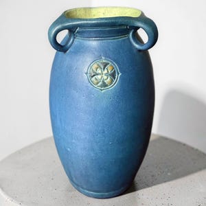 Op de afbeelding: Een matblauwe keramische vaas met twee gebogen handvatten aan de bovenkant en een cirkelvormig medaillon met een in reliëf aangebracht vierbladig bloemmotief aan de voorzijde. De binnenkant van de vaas is afgewerkt met een lichtgele glazuur.