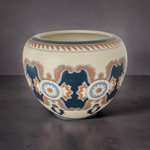 Pode incluir: Uma tigela de cerâmica creme com formato redondo. A tigela apresenta um padrão repetitivo de desenhos florais azul escuro, marrom e azul claro. A borda tem uma faixa de triângulos azuis e marrons. A tigela é provavelmente usada para fins decorativos.