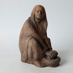 Op de afbeelding: Een terracotta beeld van een vrouw die zit met haar handen in haar schoot. Ze draagt een lange jurk en heeft lang haar. Het beeld is in zittende positie en is gemaakt van roodbruine klei.