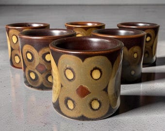 May include: A set of six cylindrical ceramic cups featuring a dark brown base color with a repeating pattern of large, muted olive-green circles and smaller, bright yellow centers. Each cup has a smooth, glazed finish and a wide, open top.