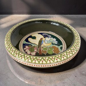 May include: A round ceramic bowl with a deep olive green interior and a wide rim featuring a repeating green and red geometric lattice pattern. The center of the bowl displays a colorful illustration of a bird perched on a branch with flowers.