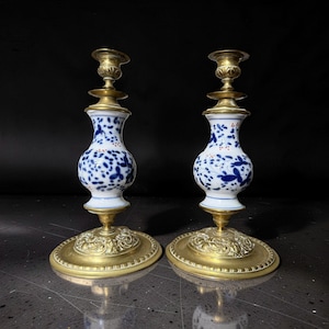 Op de afbeelding: Een paar kandelaars met witte porseleinen lichamen voorzien van blauwe bloemmotieven en kleine rode accenten. Elke kandelaar heeft een koperen voet met gedetailleerde reliëfversieringen en een bijpassende koperen kaarsenhouder.