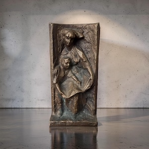 Può includere: Scultura a rilievo in bronzo raffigurante una donna e un bambino. Le figure sono in una cornice rettangolare con uno sfondo strutturato. La scultura ha una patina bronzo scuro con riflessi verdi.