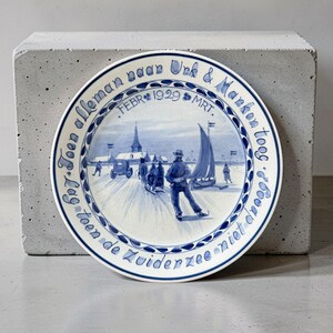 Può includere: Piatto in ceramica blu e bianco con una scena invernale di pattinatori e barche a vela. Il piatto ha un bordo blu con testo olandese, tra cui "FEBR 1929 MRT."