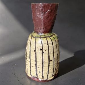 Op de afbeelding: Een keramische vaas met een hoge, donkerbruine textuurhals en een cilindrisch lichaam. Het lichaam heeft een crèmekleurig glazuur met verticale zwarte strepen en verspreide donkere spikkels, met een geribbelde oppervlaktestructuur op de basis.