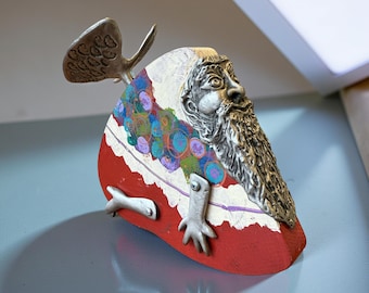 May include: A whimsical, abstract sculpture featuring a bearded face with a long beard and a wing-like element. The sculpture has a red base with white and colorful accents, and metallic arms and a wing.