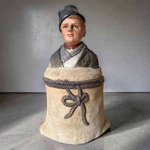 Könnte beinhalten: Eine bemalte Keramikfigur, die das Brustbild einer Person mit dunkler Mütze und Jacke darstellt, die aus einem strukturierten beigen Sack hervorkommt, der mit einem dunklen Seil verschnürt ist. Die Figur hat helle Haut, braunes Haar und einen roten Kragen.