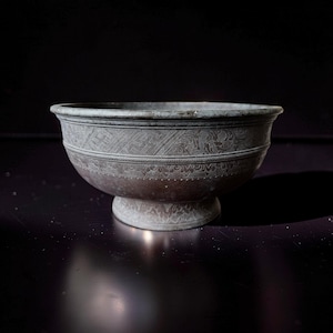 Op de afbeelding: Een gedetailleerde, antieke metalen kom met een voet. De kom heeft ingewikkelde, gegraveerde patronen rond de body, waaronder bloemen en geometrische ontwerpen. De kleur is een gedempt brons, wat een vintage uitstraling geeft.