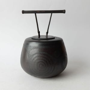 Op de afbeelding: Een zwarte keramische pot met een deksel en een metalen handvat. De pot heeft een draaiende patroon op het oppervlak.