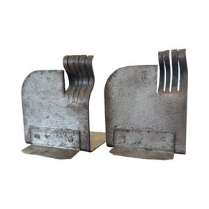 May include: Pair of industrial-style metal bookends. Each bookend has a rectangular base and a curved, textured body. One features a series of vertical, curved metal pieces, while the other has a series of vertical slits.