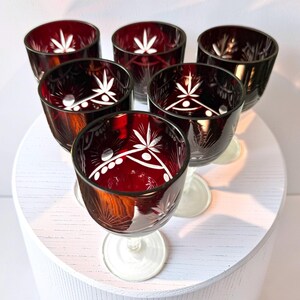 Op de afbeelding: Een set van zes dieprode glazen kelken met heldere, geslepen bloem- en geometrische patronen. Elk glas heeft een afgeronde kom, een korte steel en een ronde voet. Het glas heeft een rijke karmozijnrode kleur met gedetailleerde witte etsen.