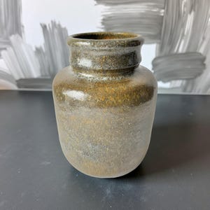 Op de afbeelding: Een handgemaakte, kleine keramische vaas met een afgeronde body en een licht uitlopende rand. De vaas heeft een gespikkeld, aards kleurenpalet met tinten bruin, goud en grijs. De buitenkant heeft een getextureerde, matte afwerking.