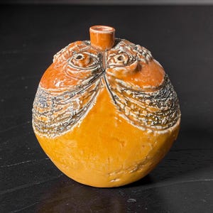 Op de afbeelding: Een ronde keramische vaas met een glanzend oranje glazuur en een kleine centrale opening aan de bovenkant. Het oppervlak is voorzien van gestructureerde, donkerbruine en crèmekleurige reliëfpatronen die rond het bovenste gedeelte lopen.
