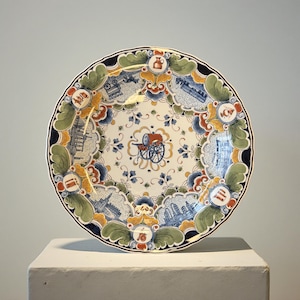 Op de afbeelding: Een decoratieve keramische plaat met een geschulpte rand. De plaat heeft een centraal ontwerp van een rode en blauwe kar, omgeven door blauwe en witte architectonische illustraties, bloempatronen en een donkerblauwe rand. De plaat wordt op een wit oppervlak weergegeven.