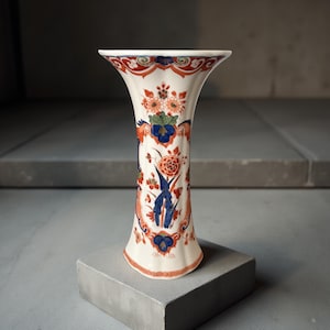Può includere: Un vaso di ceramica bianca con la parte superiore svasata e una base stretta. Il vaso è decorato con intricati motivi floreali nei toni del rosso, blu, arancione e verde. Il design è simmetrico, con motivi ripetuti. Il vaso è appoggiato su una superficie grigia.