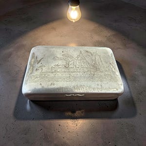 Puede incluir: Una caja rectangular de metal plateado con una imagen grabada detallada de un barco. La caja tiene una tapa con bisagras y una apariencia ligeramente desgastada. La imagen está iluminada por una bombilla colgante.