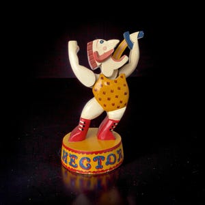 Op de afbeelding: Vintage houten beeldje van een circusartiest genaamd Hector. Het beeldje is beschilderd met een gele turnpakje, rode laarzen en een blauwe en gele hoorn. De basis is geel met de naam "HECTOR" in blauwe letters.