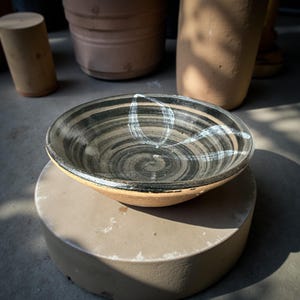 May include: A handmade ceramic bowl with a black and white spiral design. The bowl has a wide, shallow shape and a natural, earthy color. White brushstrokes add a decorative touch. The bowl is sitting on a round, light-colored pedestal.
