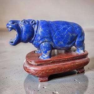 Peut inclure: Une figurine d'hippopotame sculptée en lapis-lazuli bleu, la gueule ouverte, posée sur un socle en bois brun foncé poli orné de lignes décoratives dorées sur les côtés et reposant sur quatre petits pieds.