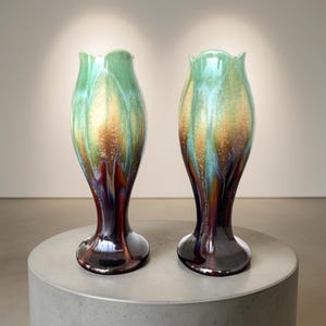 Peut inclure: Deux vases en céramique de forme tulipe. Les vases présentent une finition brillante avec des couleurs vertes, dorées, bleues et bordeaux. Ils sont posés sur un socle rond gris.