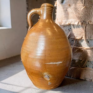 Puede incluir: Una jarra de cerámica alta y bulbosa con un esmalte brillante de color marrón miel. El recipiente cuenta con una sola asa curva unida a un cuello estrecho con crestas horizontales. La superficie muestra variaciones sutiles de color y una marca.