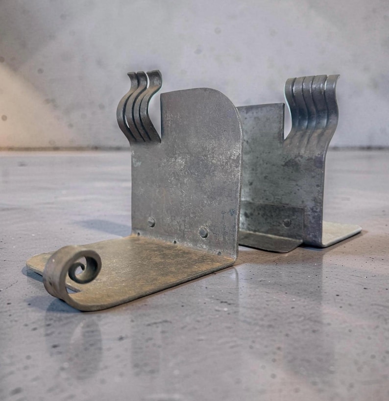 May include: A pair of silver metal bookends. Each bookend has a rectangular base and a vertical support with a decorative, curved design. One bookend features a scroll detail on the base. The metal has a slightly weathered appearance.
