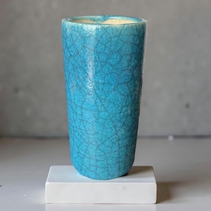Könnte beinhalten: Eine hohe, zylindrische türkisfarbene Keramikvase mit einem Craquelé-Effekt. Die Vase steht auf einem kleinen, weißen quadratischen Sockel. Das Innere der Vase ist hellcremefarben. Die Vase ist ein dekorativer Gegenstand.