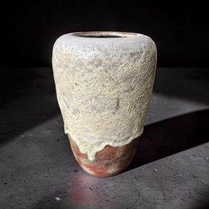 Può includere: Un vaso in ceramica fatto a mano con una parte superiore strutturata bianco sporco e una base marrone-rossastra. Il vaso ha una forma organica unica e un effetto smaltato a goccia, alto circa 20 cm.