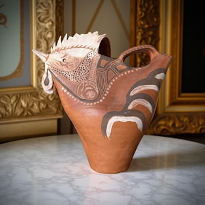 Op de afbeelding: Terracotta keramische kan met een hanenontwerp. De kop en kam van de haan zijn wit, met bruine en zwarte decoratieve patronen. De kan heeft een handvat en staat op een marmeren oppervlak.