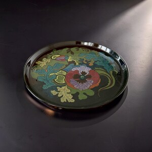 May include: A round, dark-colored decorative tray featuring a central floral motif with a large, multi-colored pansy in shades of purple, red, and orange, surrounded by stylized green and teal leaves and intricate gold-colored line details.