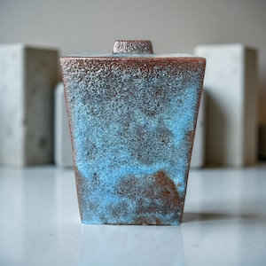Peut inclure: Un vase carré en céramique à surface texturée. Le vase a un dessus et une base marron, avec un dégradé bleu et marron au milieu. Le vase a un petit dessus cylindrique et est placé sur un fond flou.