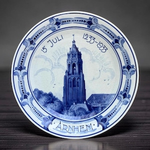 May include: A round ceramic plate featuring a blue and white hand-painted illustration of a tall church tower in Arnhem. The text 15 JULI 1233-1933 is printed at the top, and the word ARNHEM is printed at the bottom within a decorative border.