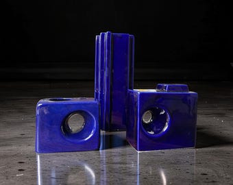 May include: Three deep cobalt blue ceramic objects with a glossy finish. One tall, rectangular piece with vertical ridges stands behind two shorter, square-shaped pieces, each featuring a circular opening on the front face and a flat top surface.
