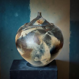Op de afbeelding: Een handgemaakte keramische pot met deksel. De ronde pot heeft een donkere, gespikkelde glazuur in bruin, zwart en crème. Hij staat op een donkere vierkante sokkel tegen een blauw-beige achtergrond.