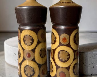 May include: A pair of cylindrical ceramic salt and pepper shakers featuring a dark brown base color with a repeating pattern of large yellow circles, smaller tan floral motifs, and orange dots, topped with a smooth, light brown conical cap.