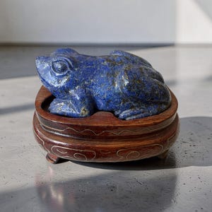 Op de afbeelding: Een uit lapis lazuli gesneden kikkerfiguur met een diepblauwe kleur en goudkleurige spikkels, rustend op een donkerbruine houten voet met dunne, lichtgekleurde decoratieve draadinlegpatronen langs de gebogen, verhoogde zijkanten.