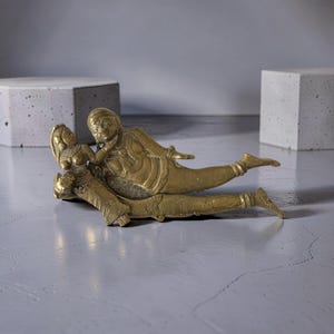 Op de afbeelding: Een gedetailleerde messing sculptuur die twee figuren in een liggende pose afbeeldt. De figuren zijn versierd met ingewikkelde details, die traditionele kleding en versieringen laten zien. De sculptuur heeft een warme, gouden tint en staat tegen een neutrale achtergrond.