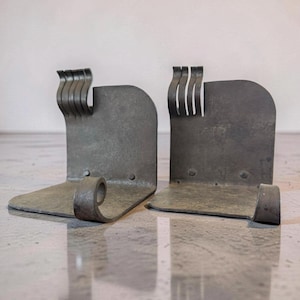 May include: A pair of metal bookends with a rustic, industrial design. Each bookend has a curved base, a vertical back, and a decorative, wavy top. The metal has a weathered, gray finish, adding to the vintage aesthetic.