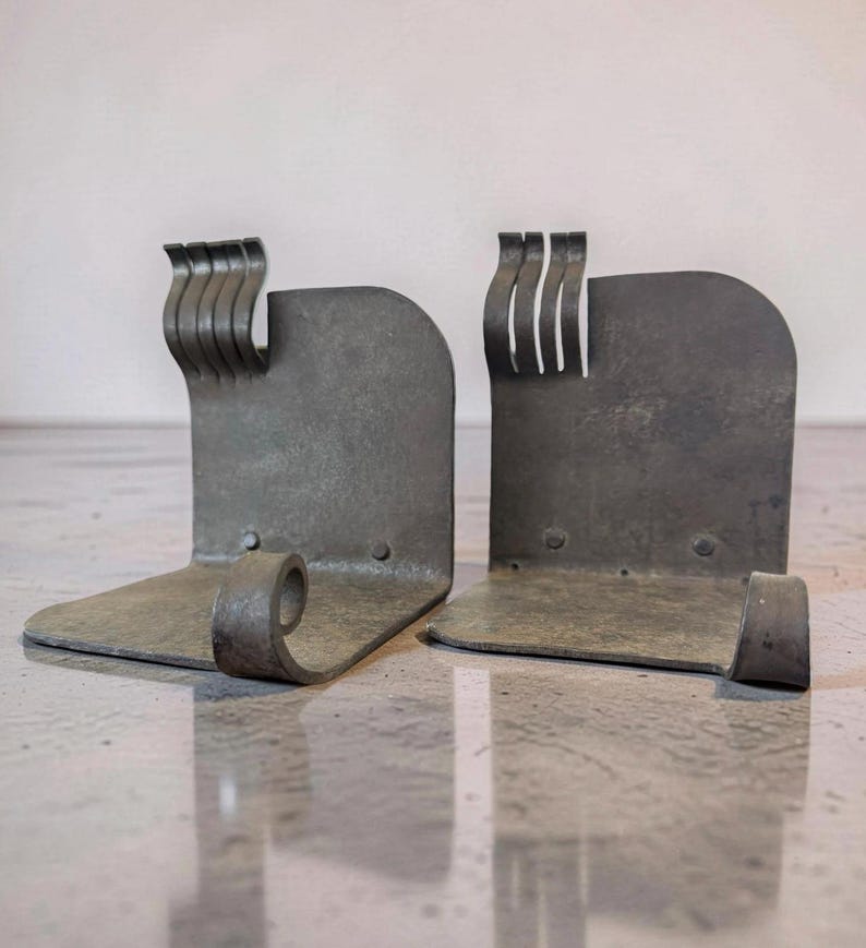 May include: A pair of metal bookends with a rustic, industrial design. Each bookend has a curved base, a vertical back, and a decorative, wavy top. The metal has a weathered, gray finish, adding to the vintage aesthetic.